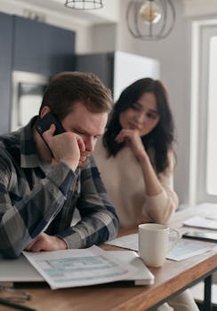 A worried couple sits at a table with bills and a phone, planning their finances.