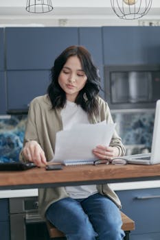 A young woman reviews documents and uses her laptop in a modern kitchen setting.