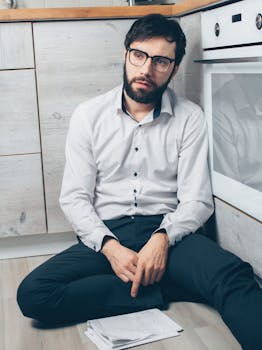 Distressed young man sitting in a kitchen, looking worried with papers on the floor.